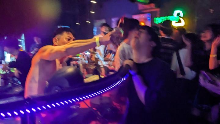 Shirtless man behind the counter holding a bottle of hard liquor over the mouth of a another person on the other side