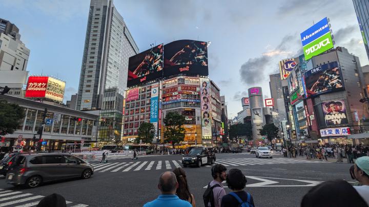 The image captures the famous Shibuya Crossing in Tokyo, Japan, during the evening. The scene is filled with pedestrians waiting to cross the wide, zebra-striped intersection while vehicles pass through. Surrounding the crossing are towering buildings adorned with bright, colorful billboards, neon signs, and digital advertisements, including recognizable brands like Uniqlo, H&M, and Joysound. The atmosphere is bustling, with a mix of locals and tourists. The sky is transitioning from daylight to dusk, adding to the vibrant cityscape.