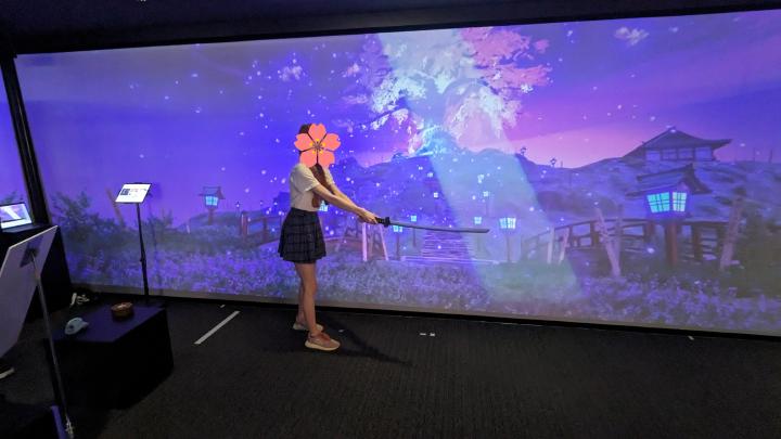 Yuu is swinging a prop sword. On the wall behind her, the image of Saigyou Ayakashi is displayed with a flare following the sword movement